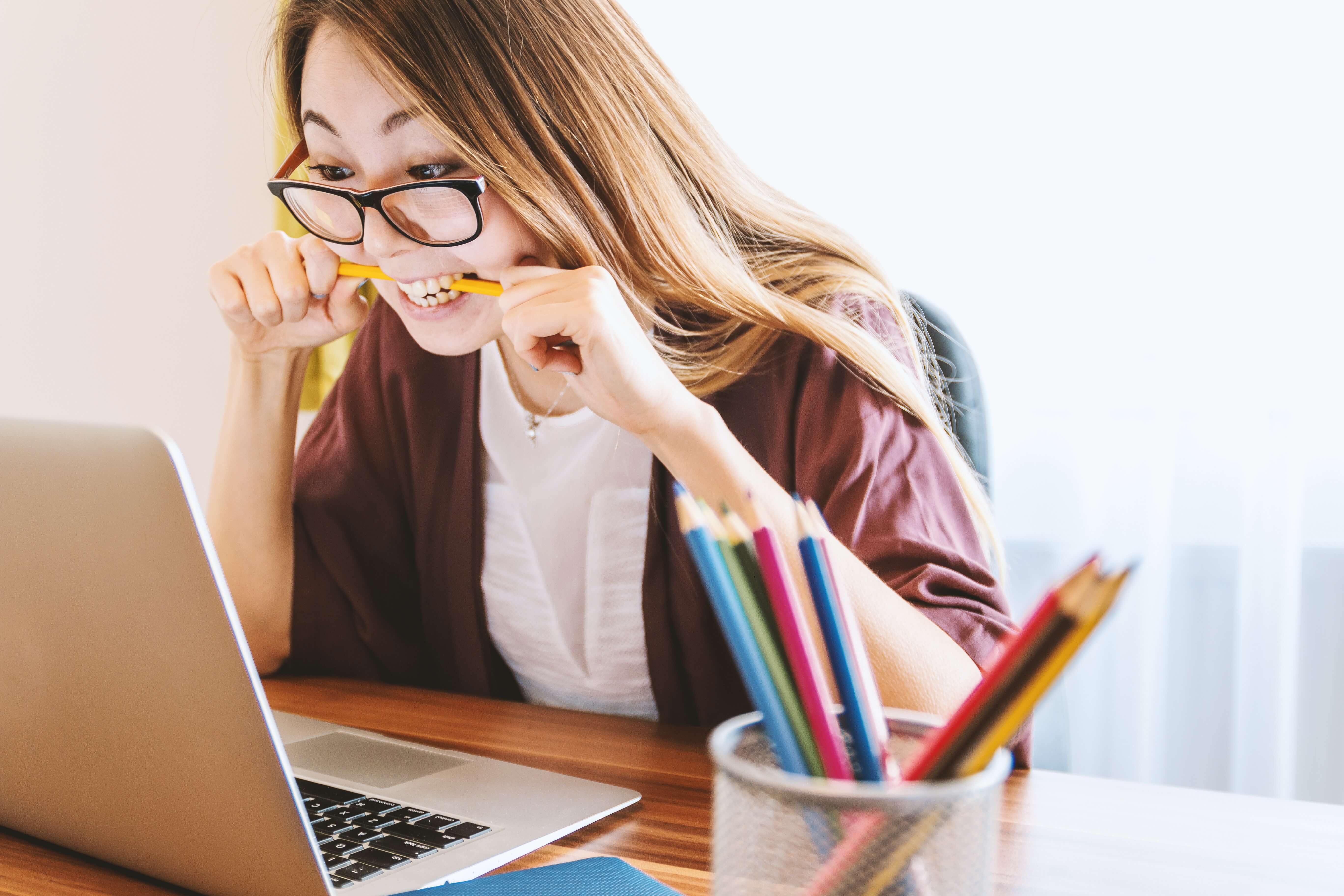 A woman looking at her laptop screen and biting a pencil in frustration.